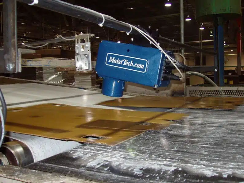 A machine with a "MoistTech" sensor is positioned above moving materials on a conveyor belt in a factory setting.